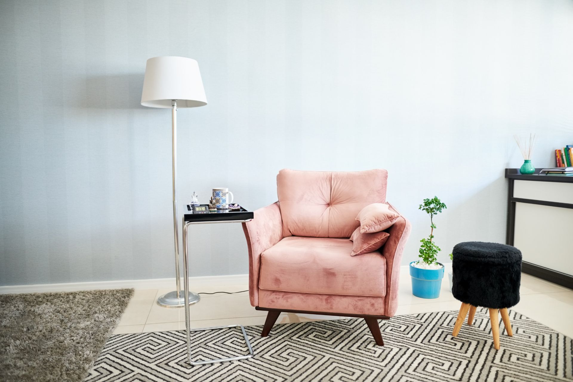A living room scene with a pink armchair, side table with a mug and tray, floor lamp, black footstool, blue potted plant, and geometric patterned rug.